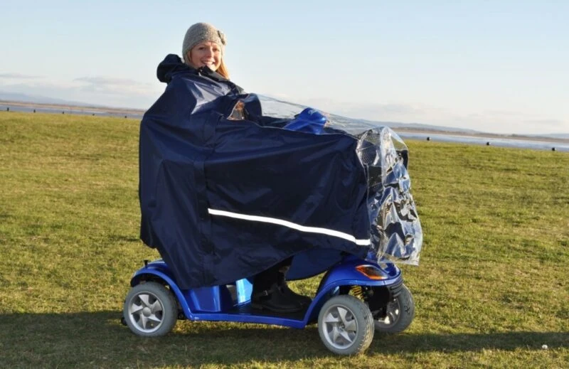 Person using a mobility scooter with a protective cape in an open field.