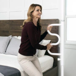 Woman using a white curved grab bar next to a bed in a bedroom.