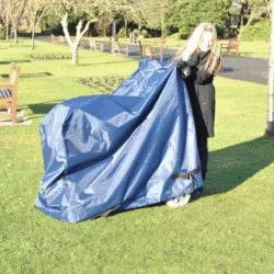 Person covering a mobility scooter with a navy waterproof cover in a park.