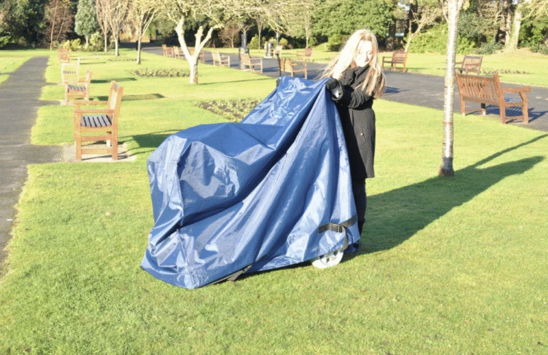 Person covering a mobility scooter with a navy waterproof cover in a park.