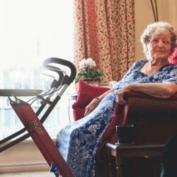 Elderly woman seated next to a red indoor rollator with a tray.