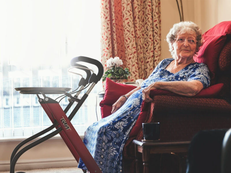 Elderly woman seated next to a red indoor rollator with a tray.