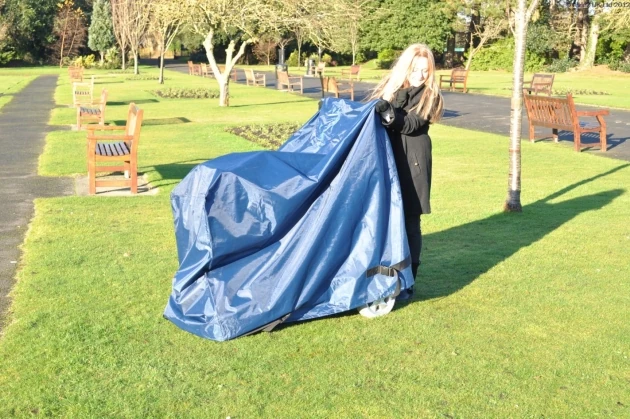 Person covering a mobility scooter with a navy waterproof cover in a park.