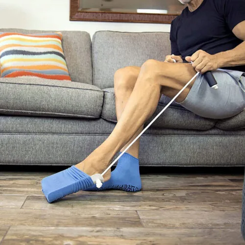 Person using a sock stocking aid to put on blue socks while seated on a couch.