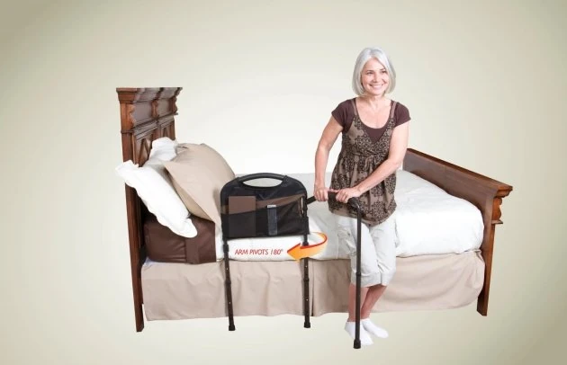 Woman using a mobility bed rail with a swing-out arm next to a wooden bed.