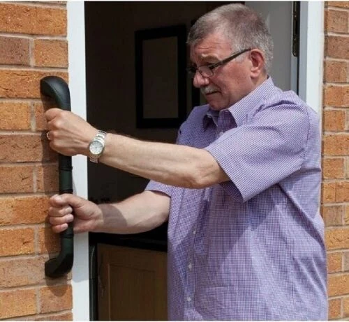 Elderly man gripping a black outdoor safety handle on a brick wall.