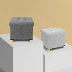 Two pouffe stools with storage in light and dark grey on display blocks.