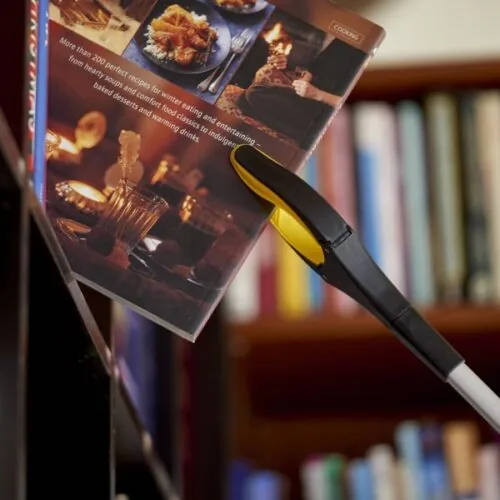 Helping hand grabber holding a book near a bookshelf.