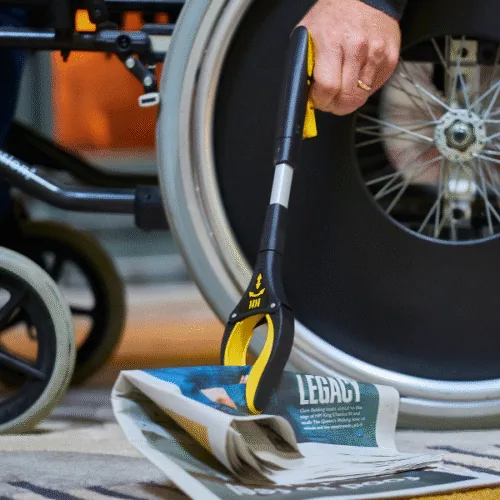 Person using a small grabber tool to pick up a newspaper near a wheelchair.