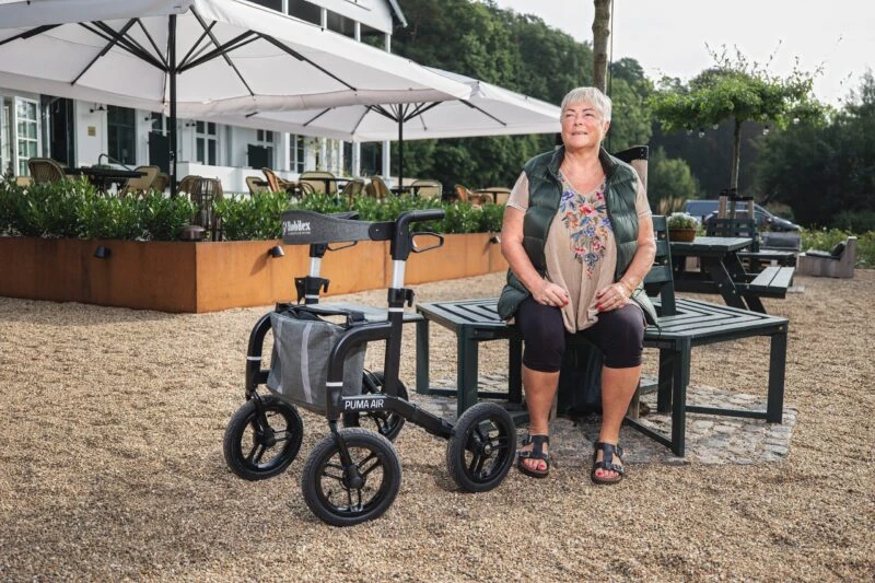 Woman sitting on a bench next to a Mobilex Puma Air rollator with large wheels.