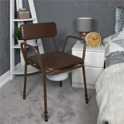 Brown commode chair with padded back next to a bedside table and bed.