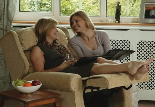 Two women sitting on a beige recliner chair, smiling and talking.