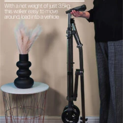 Person holding a folded walking frame with wheels beside a small table.