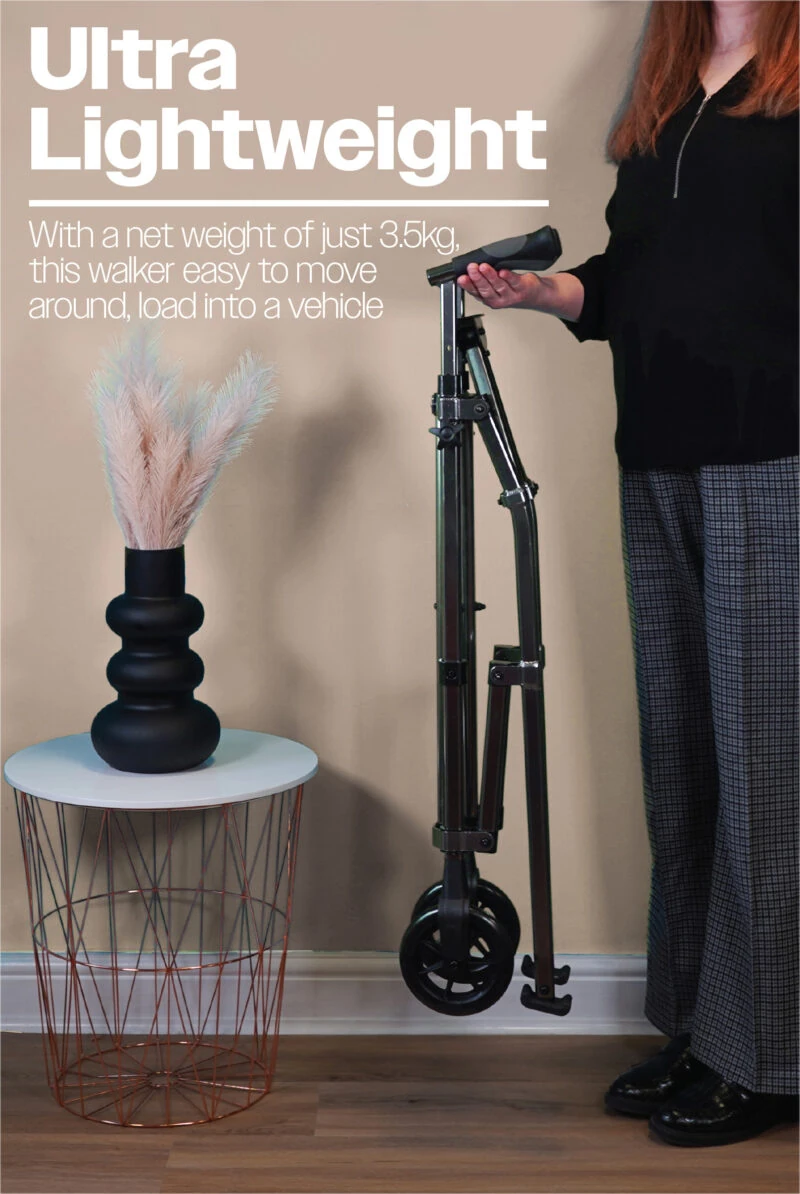 Person holding a folded walking frame with wheels beside a small table.