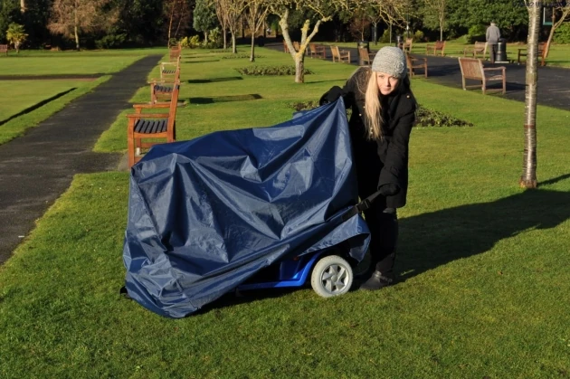 Person covering a mobility scooter with a navy waterproof cover in a park.