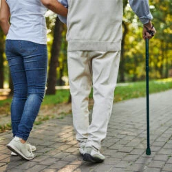 Person using a walking stick while walking on a paved path with a companion.