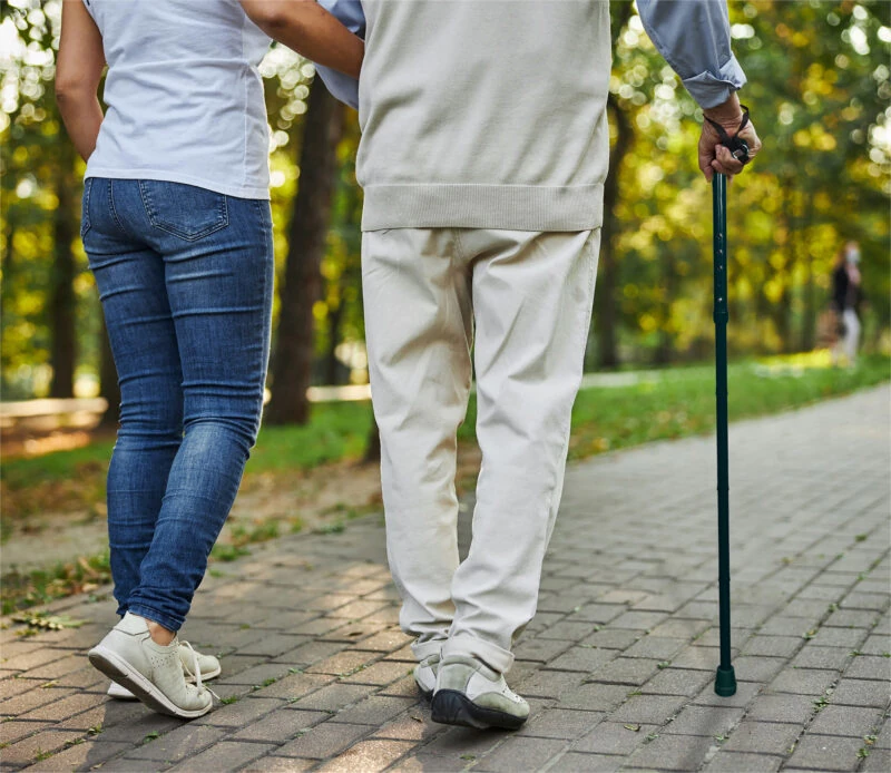 Person using a walking stick while walking on a paved path with a companion.