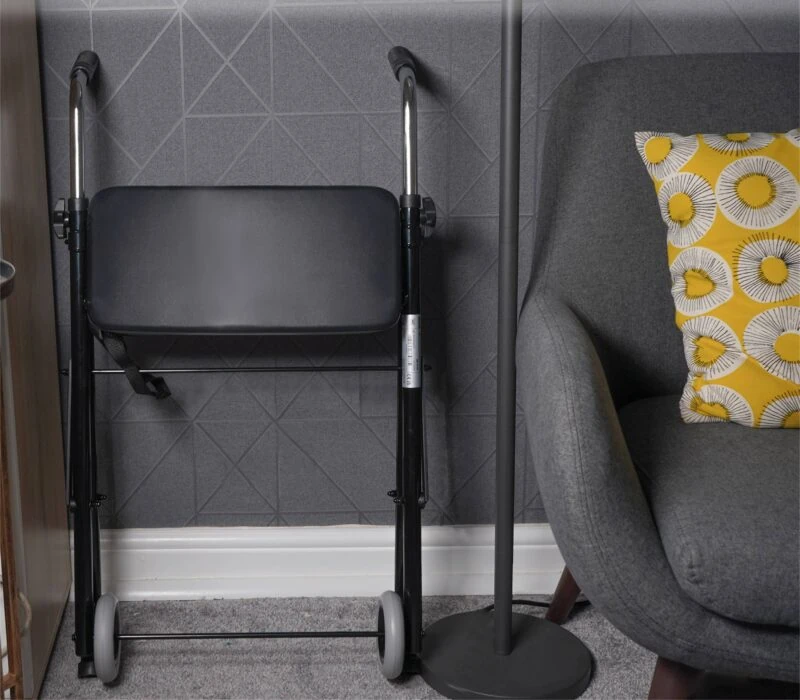 Folded wheeled walker with seat next to a chair and lamp.