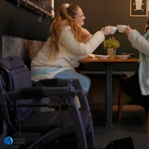 Rollz Motion Electric Rollator parked beside a cafe table with two people enjoying drinks.