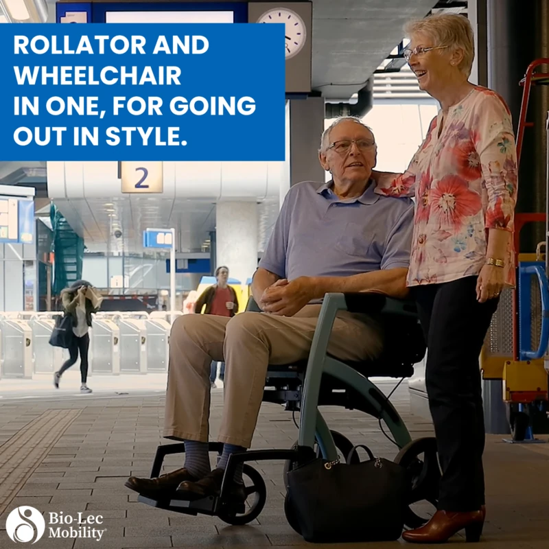 Elderly man seated on Rollator Wheelchair with woman standing beside him at a station.