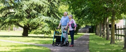Two people walking with a Rollator Wheelchair on a tree-lined path.
