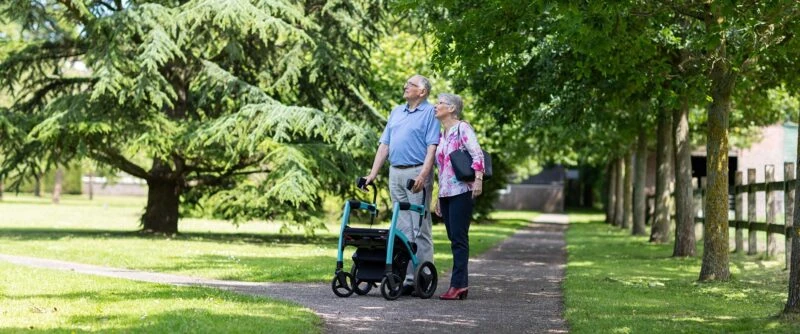 Two people walking with a Rollator Wheelchair on a tree-lined path.