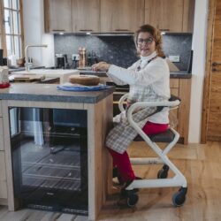 Person seated on Saljol Indoor Rollator with a wooden seat in a kitchen.