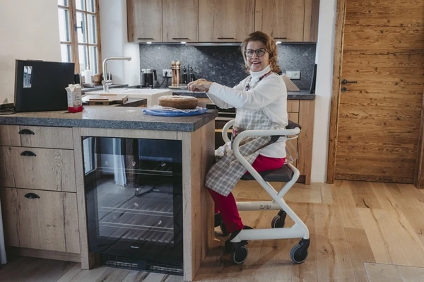 Person seated on Saljol Indoor Rollator with a wooden seat in a kitchen.