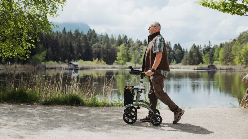 Man walking with a rollator near a scenic lake and forest.