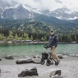 Person using an off-road rollator on a rocky lakeside path with mountains in the background.