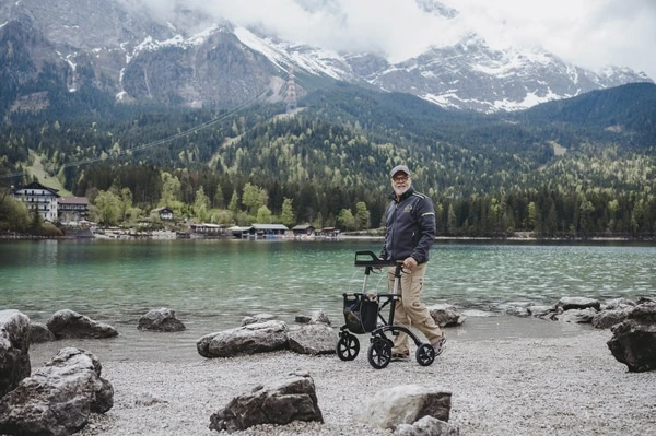 Person using an off-road rollator on a rocky lakeside path with mountains in the background.