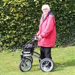 Person using Trionic 14er Rollator Walker on grass with a hedge background.