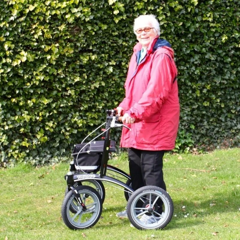 Person using Trionic 14er Rollator Walker on grass with a hedge background.