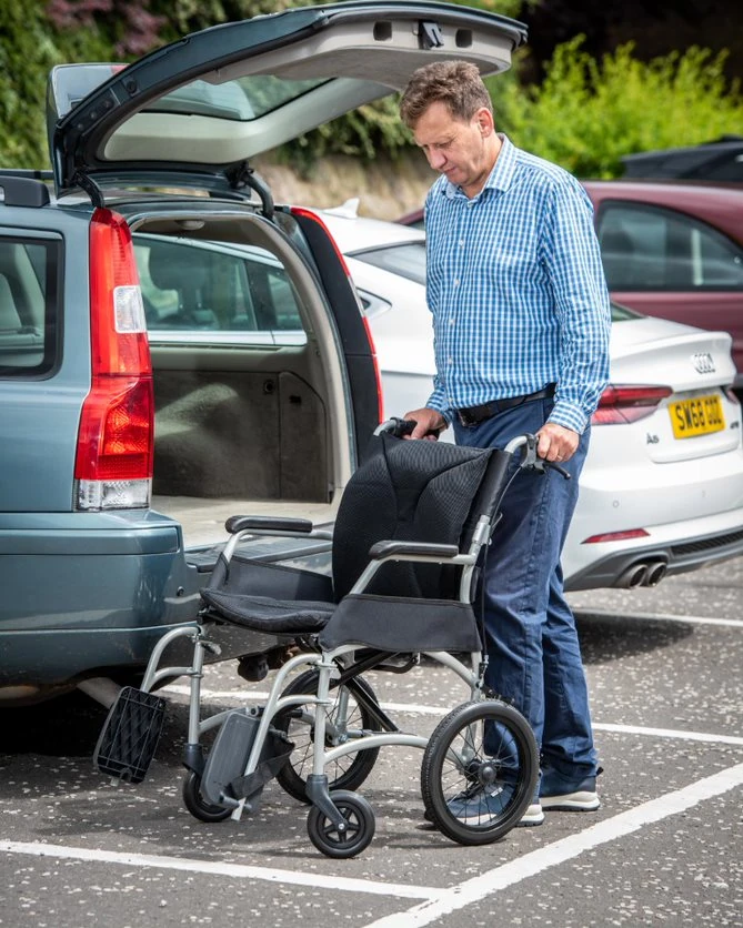 Man standing next to a lightweight folding wheelchair by a car.