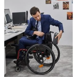 Person in a suit sitting in a compact power wheelchair at an office desk.