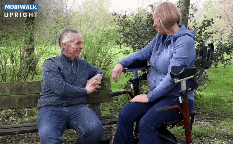 Two people sitting, one using a MobiWalk Upright Walker with pneumatic wheels.