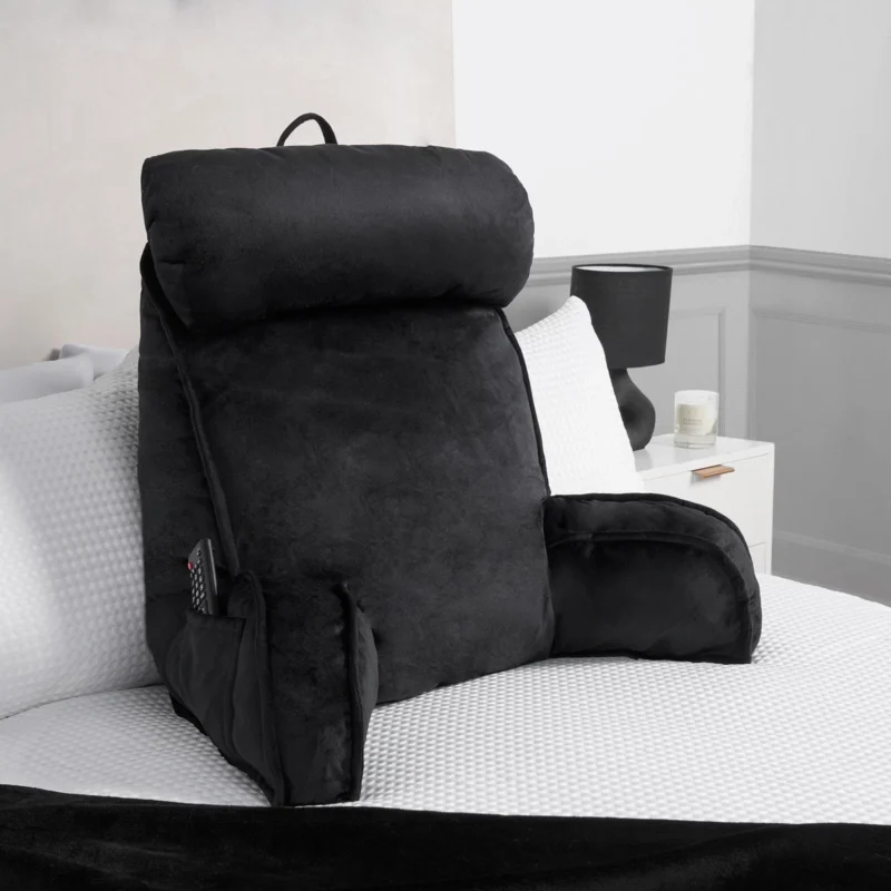 Black lumbar support pillow on a bed with a remote in the side pocket.